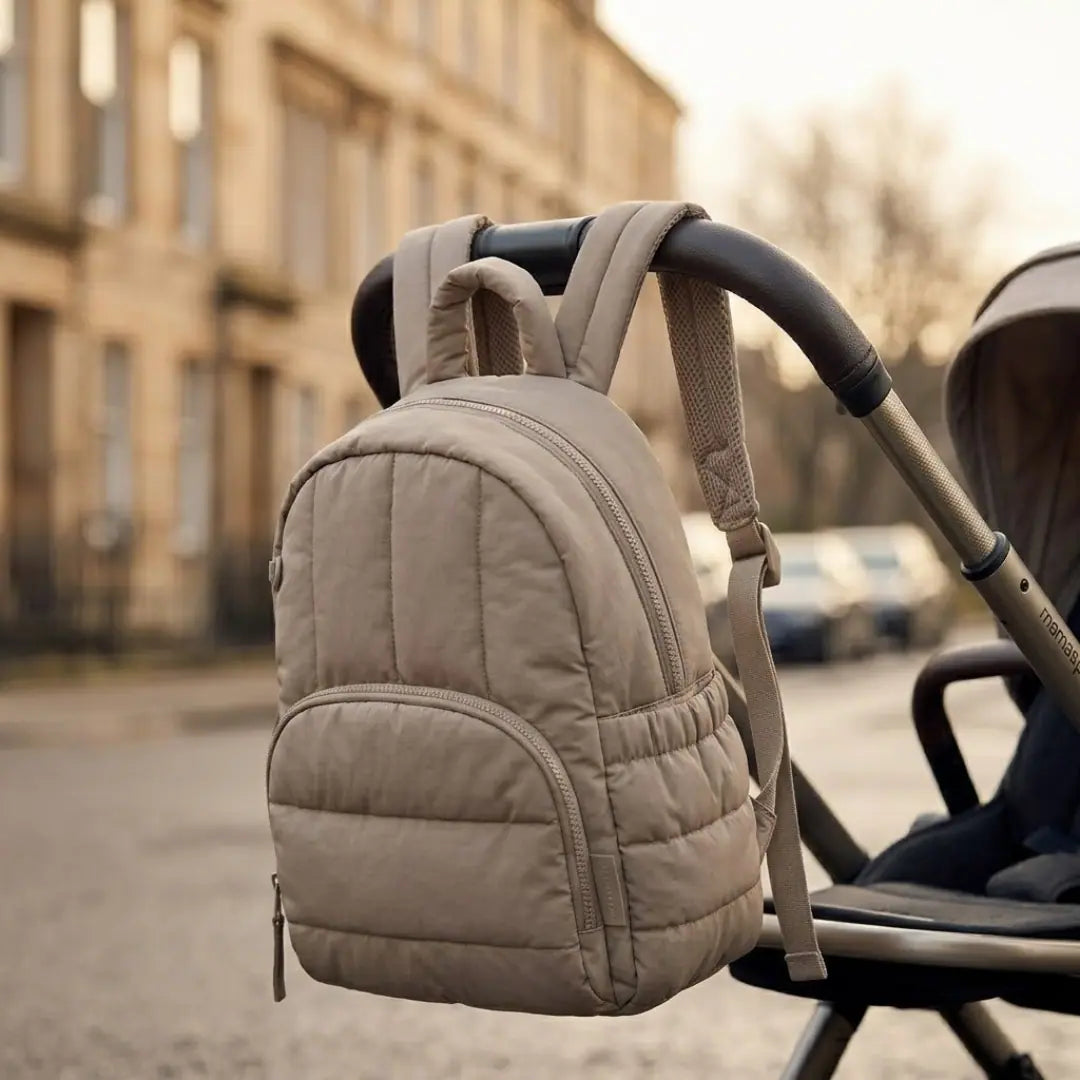 Petit sac à langer beige matelassé accroché à la poignée d'une poussette, dans une rue en ville, format compact pour une sortie légère